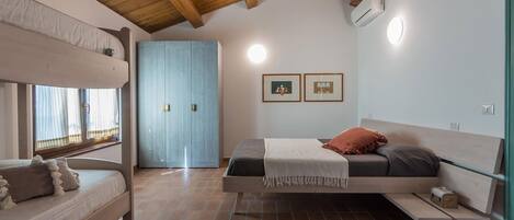 A bedroom with a bunk bed, a single bed, a wooden ceiling, a window with curtains, and two framed pictures on the wall.