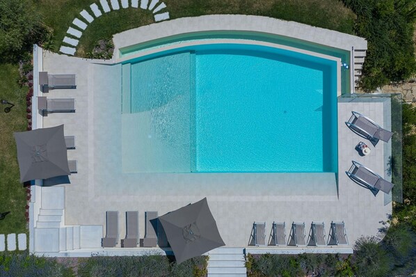 Vista aérea de una piscina con borde curvo, rodeada de sillas de descanso y sombrillas.