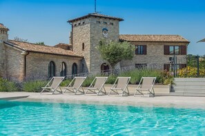 Zona de piscina con sillones de descanso y un edificio de piedra con torre.