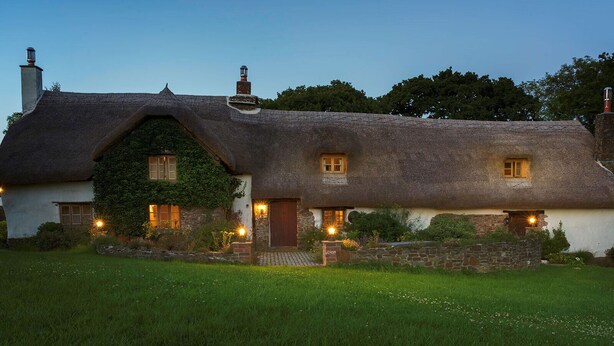 A thatched-roof cottage with a stone foundation, ivy-covered walls, and multiple windows with warm lighting.