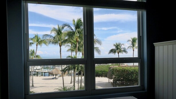 Una habitación con un amplio ventanal con vista a una playa con palmeras y cielo despejado.