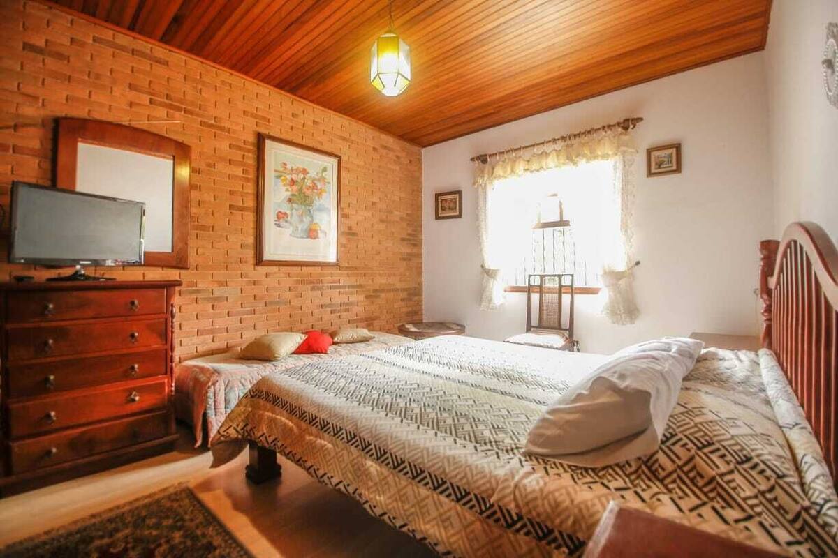 A bedroom with a brick wall, wooden ceiling, a bed with patterned bedding, a dresser, a television, and a window with curtains.
