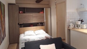 Una habitación compacta con cama, un pequeño sofá, un armario de madera y una kitchenette con lavabo y cafetera.