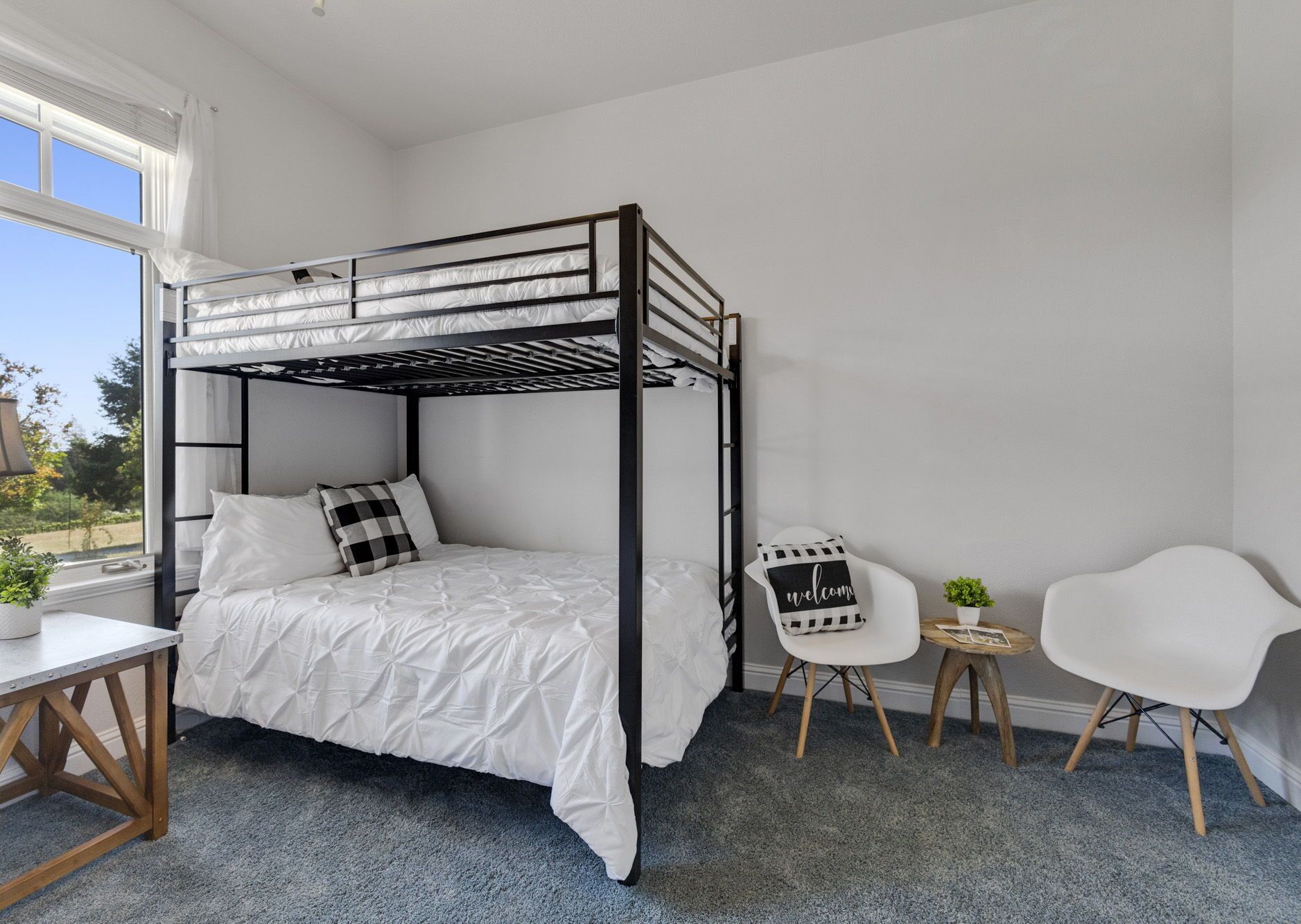 Un lit superposé avec un matelas blanc et des oreillers noirs et blancs, une petite chaise blanche, une table ronde en bois et une petite plante en pot.