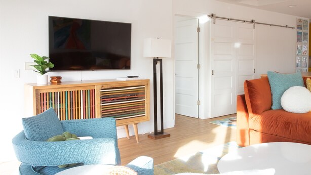 A modern living room with a flat-screen TV, a wooden console, a blue armchair, an orange sofa, and a floor lamp.