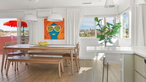Uma sala de jantar luminosa com mesa de madeira, cadeiras e luminárias pendentes. Um quadro colorido está na parede e uma planta no parapeito da janela.