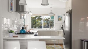 Uma cozinha moderna com armários brancos, uma geladeira de aço inoxidável e uma janela grande com vista para palmeiras.