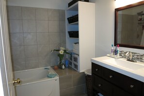 Uma casa de banho com banheira, toucador branco com espelho e prateleira com artigos de higiene pessoal.