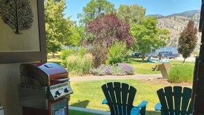Un patio couvert équipé d’un barbecue, de chaises d’extérieur et offrant une vue sur un jardin aménagé et un espace de stationnement.
