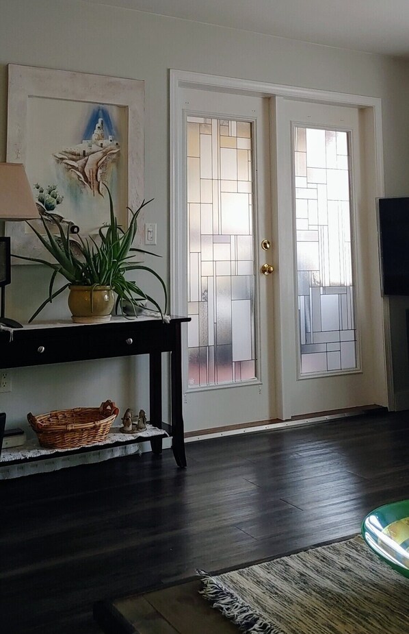 A room with a dark wooden floor, a small table with a lamp and a plant, a wall-mounted TV, and a door with frosted glass panels.