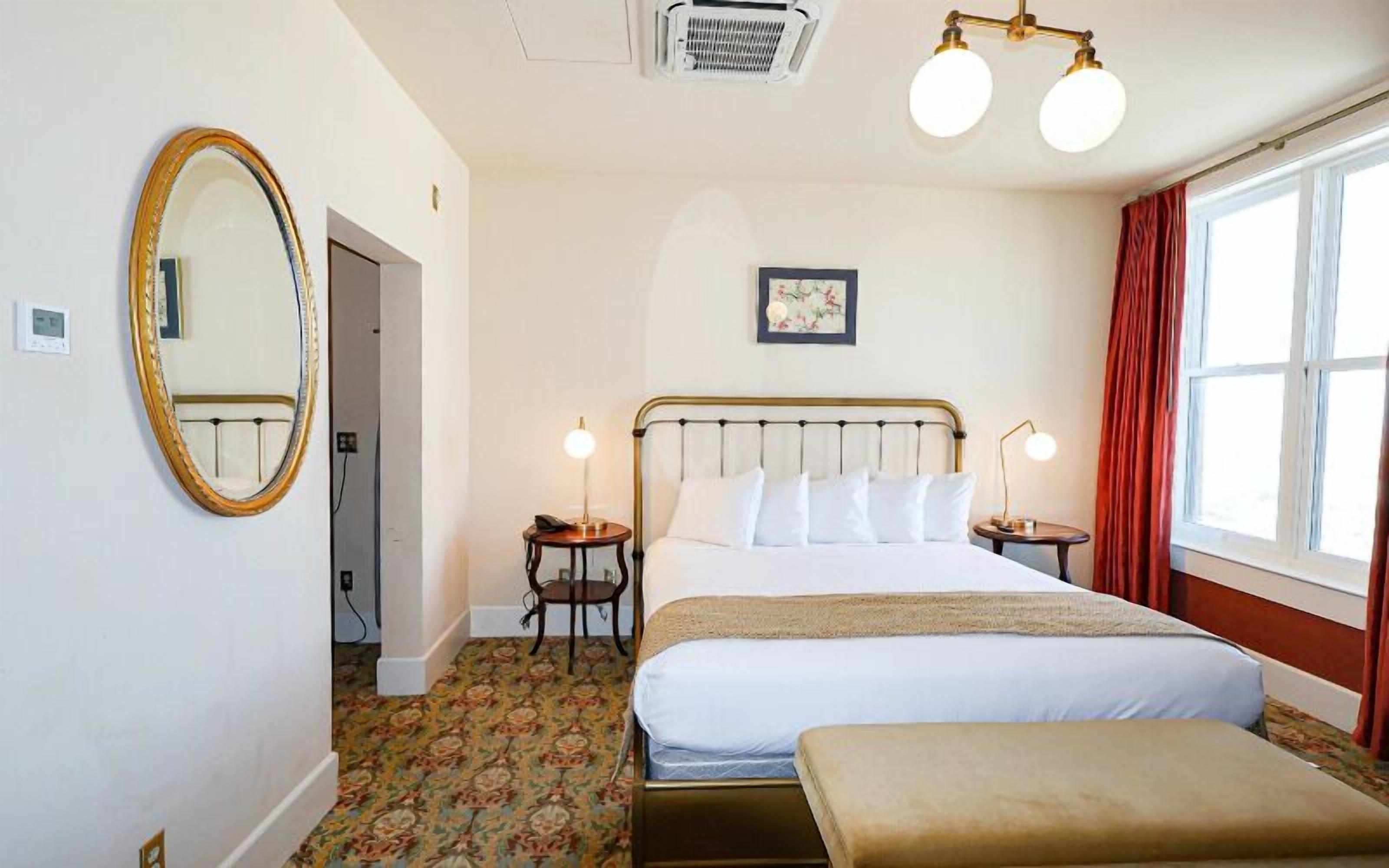A hotel room with a bed, bedside tables, a mirror, a window with curtains, and a ceiling light fixture.
