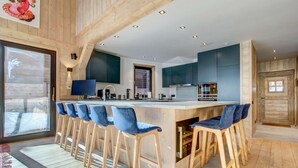 A modern kitchen with bar stools, a large island, and a view through a sliding door. - Nosefosa - Stunning new chalet with inside swimming pool (MORZINE)