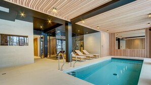 A modern indoor swimming pool with clear glass walls, lounge chairs, and a wooden ceiling design. - Nosefosa - Stunning new chalet with inside swimming pool (MORZINE)