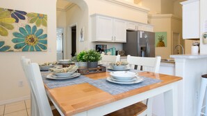 Un coin repas avec une table en bois dressée pour quatre personnes, des chaises blanches et, au fond, une cuisine avec des meubles blancs et un réfrigérateur.