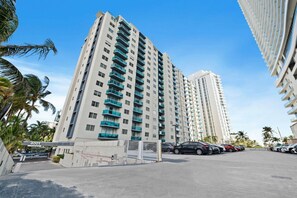 A high-rise building with multiple balconies, a gated entrance, and a parking area with several cars.