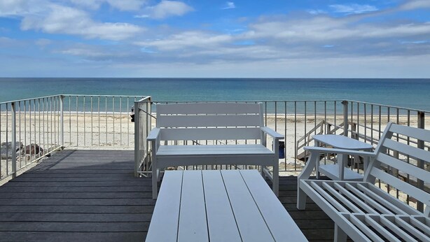 Terraza de madera con bancos y mesas blancas con vista a la playa.