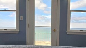 A room with a door leading to a balcony with a beach view.