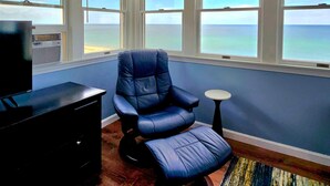 Una habitación con una silla azul, una mesita redonda pequeña, un televisor y vistas al océano a través de amplias ventanas.