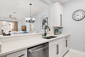 Una cocina con encimera blanca, un fregadero, lavavajillas, cafetera y un reloj en la pared.