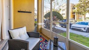 A balcony with wicker furniture, a glass table, and a view of parked cars and buildings.
