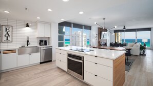 Une cuisine moderne avec des meubles blancs, un four encastré et un grand îlot central. Un coin repas offre une vue sur l’océan grâce à de grandes fenêtres.