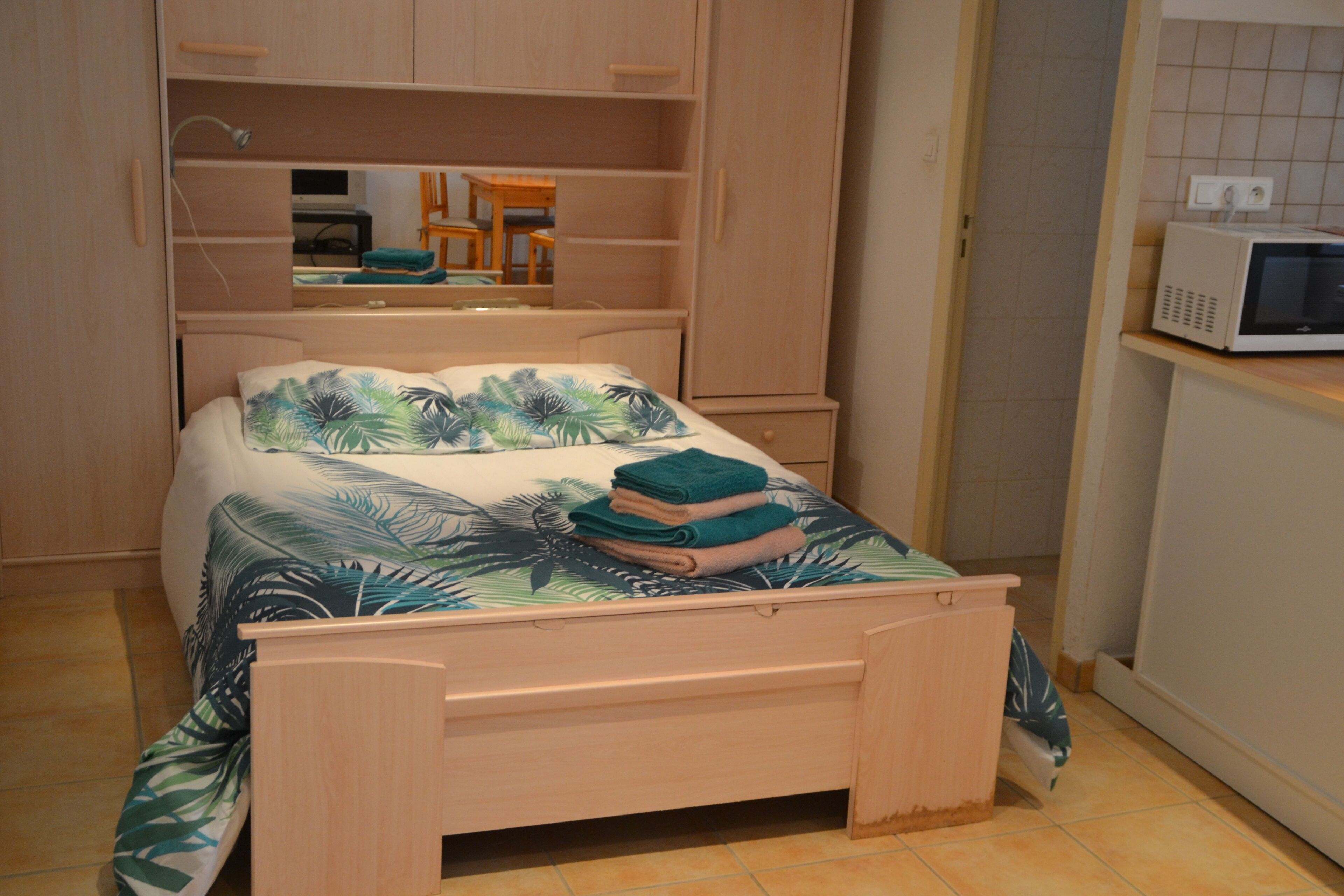 Een slaapkamer met een bed, een houten garderobe, een spiegel, een magnetron en een klein tafeltje met stoelen.