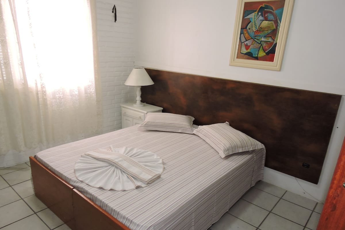 A neatly made bed with striped bedding and pillows, a bedside lamp, and a framed abstract painting on the wall.