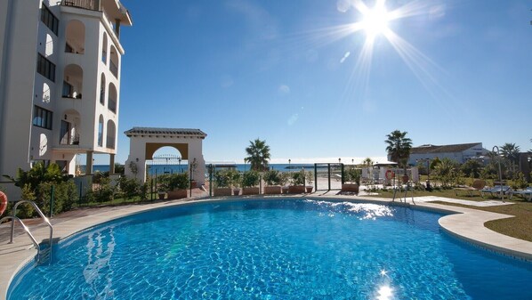 A swimming pool with a clear view of the sea, a building with balconies, and a palm tree.
