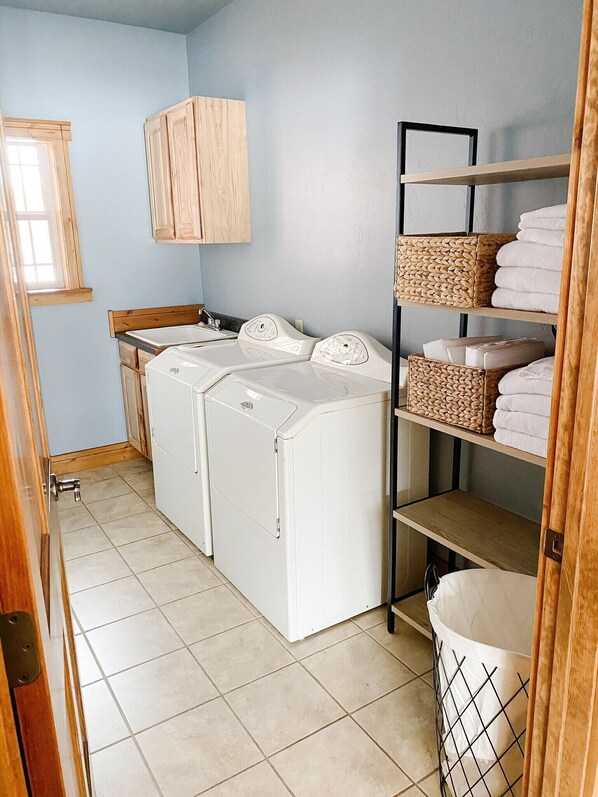 A laundry room with two front-loading washing machines, a wooden cabinet, a shelf with folded towels, and a trash bin.