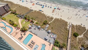 Luftaufnahme eines Strandresorts mit Schwimmbecken, Liegestühlen und einer Promenade, die zum Strand führt.