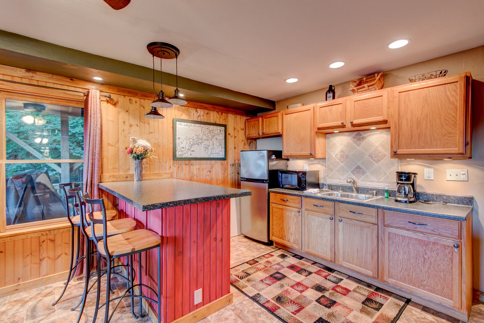 Cocina con armarios de madera, refrigerador de acero inoxidable, microondas y cafetera. Cuenta con una barra con taburetes y vistas al exterior a través de una ventana.