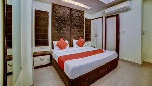 A bedroom with a large bed, a wooden headboard, a bedside table, a wardrobe, and an air conditioning unit. - Cosmo Hotel (New Delhi)