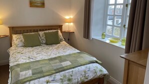 A neatly made bed with a floral duvet, two bedside lamps, a window with curtains, and a small table beside the window.