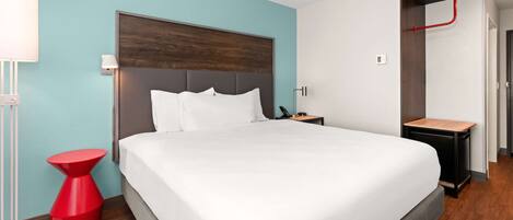 Una habitación de hotel moderna con una cama grande, una mesita de noche roja de acento, una lámpara de noche y una mesita auxiliar de madera.