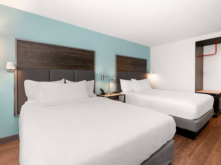 A hotel room with two beds, a nightstand, a lamp, and a red pipe on the ceiling.
