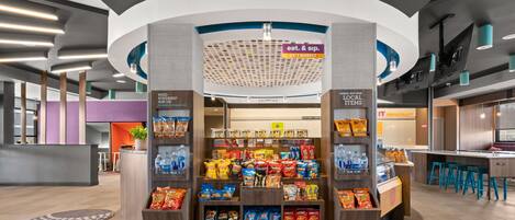 A snack kiosk with a variety of packaged snacks and beverages, a sign indicating a 24/7 market, and a checkout area with a counter and stools.