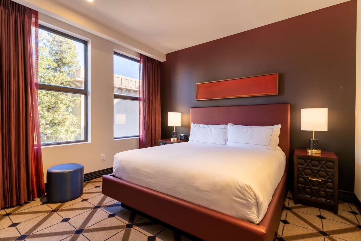 A hotel room with a large bed, two bedside tables with lamps, a small round ottoman, and a window with curtains.