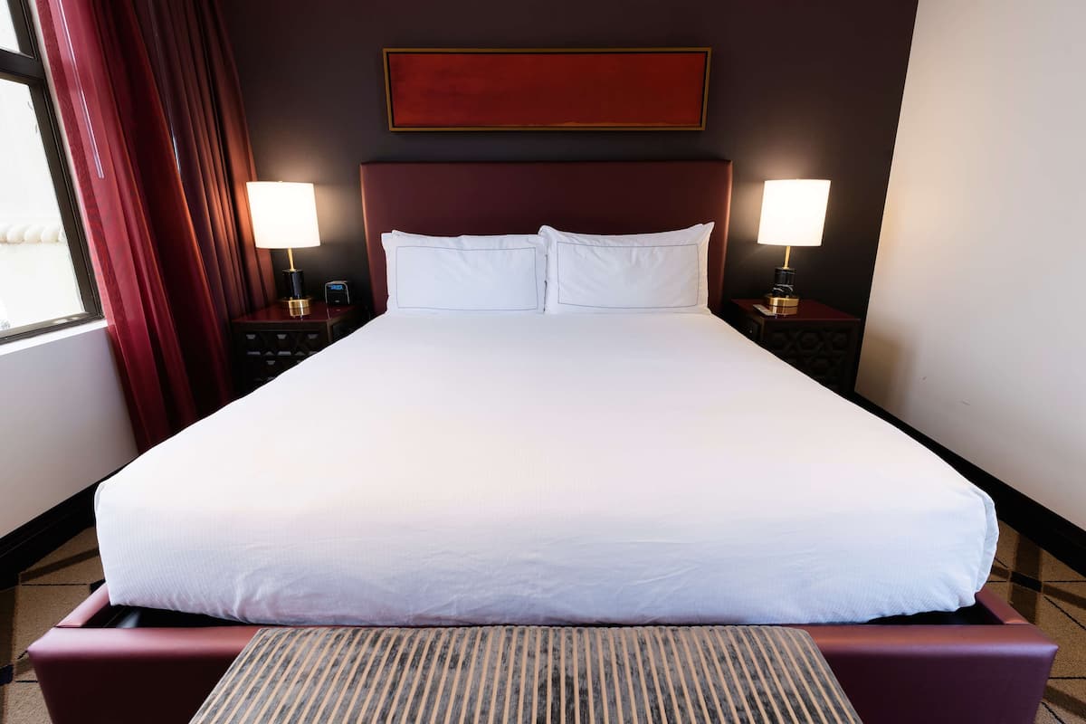 A large bed with white linens, two bedside tables with lamps, a dark headboard, and a window with red curtains.