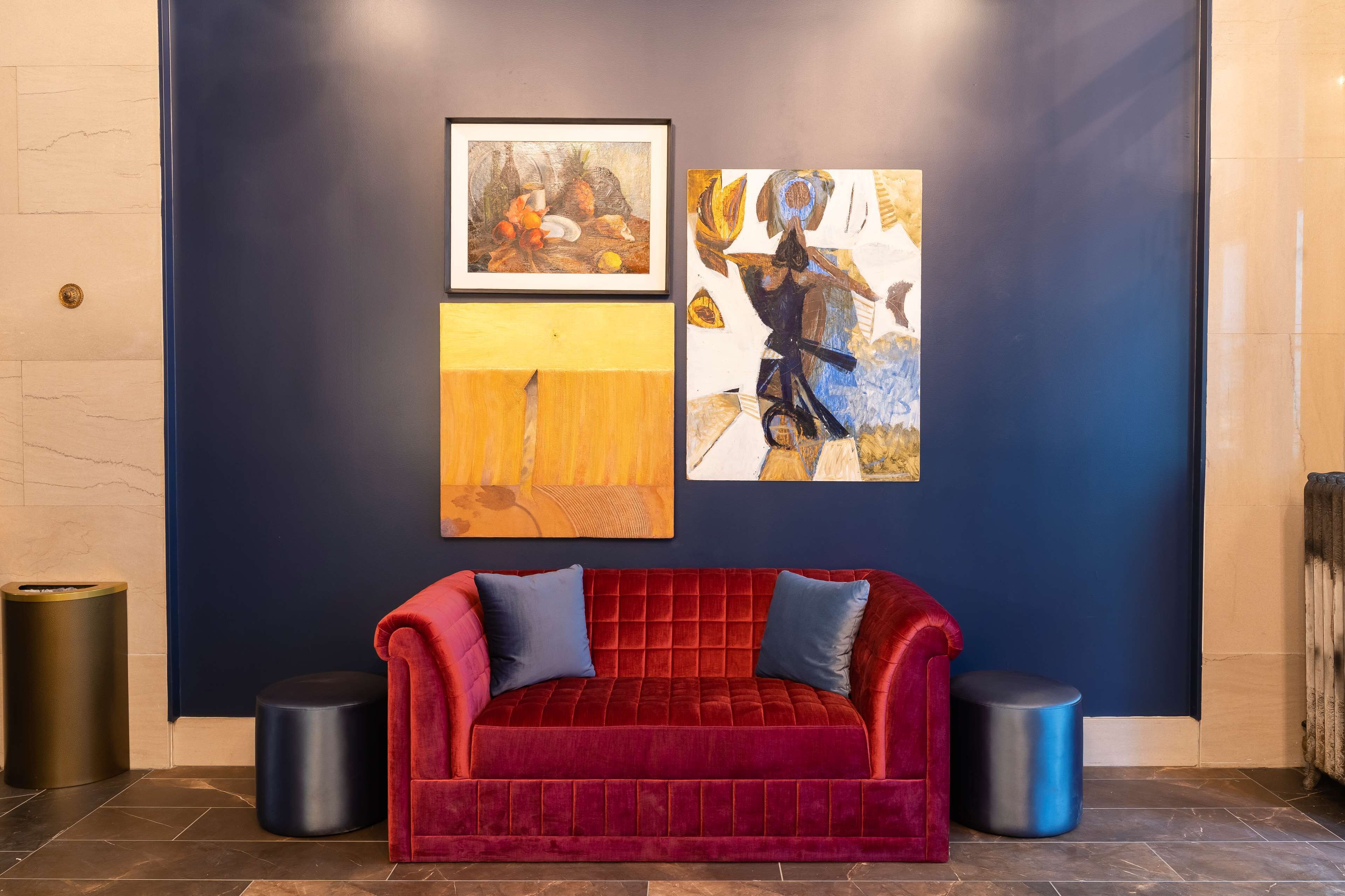 A red velvet sofa with blue cushions, a framed painting of fruits, and another abstract painting on a dark blue wall.