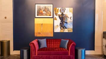 A red velvet sofa with blue cushions, a framed painting of fruits, and another abstract painting on a dark blue wall.