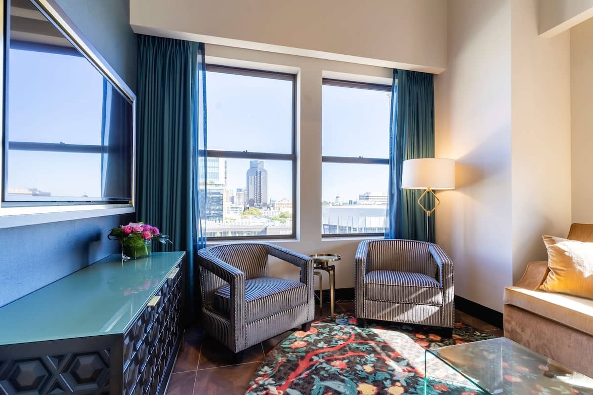A hotel room with two large windows, a sofa, a chair, a small table, and a decorative rug.