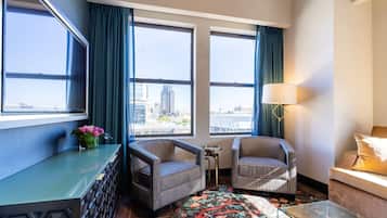 A hotel room with two large windows, a sofa, a chair, a small table, and a decorative rug.