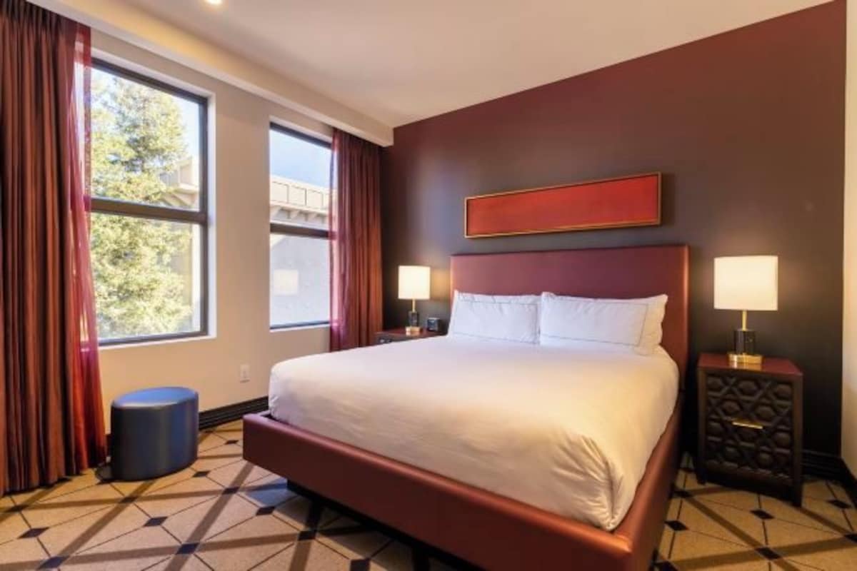 A hotel room with a large bed, two bedside tables with lamps, a window with curtains, and a geometric patterned floor.