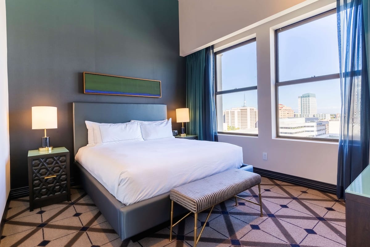 A hotel room with a large bed, two bedside tables with lamps, a bench, and a city view through large windows.