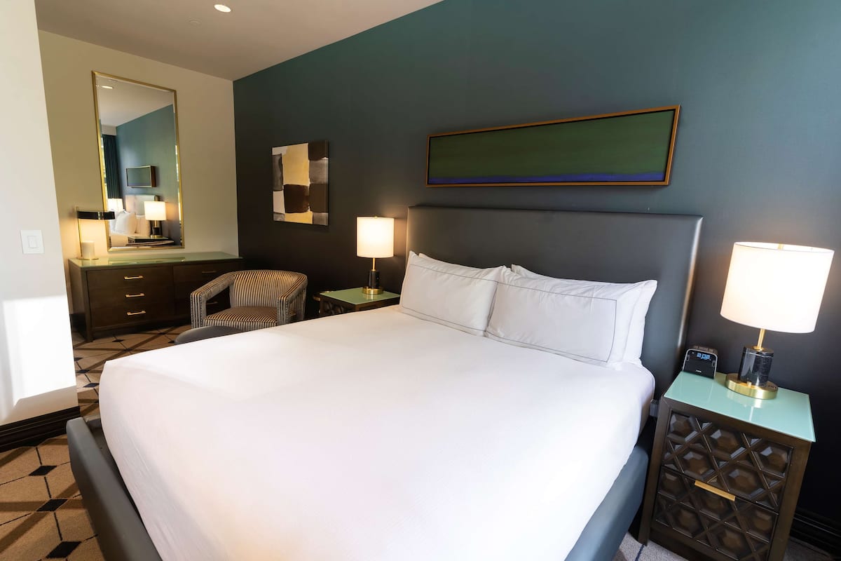 A hotel room with a large bed, a desk with a chair, a nightstand with a lamp, and a wall-mounted painting.