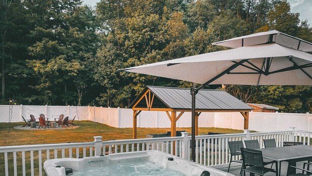 Un bain à remous sur une terrasse en bois, agrémentée d’une pergola et d’un parasol, le tout entouré d’une clôture blanche et d’arbres.