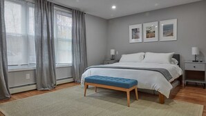 Une chambre à coucher avec un grand lit, un banc, une table de chevet, une lampe fixée au mur et trois tableaux encadrés.