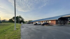 A motel with a parking lot, a covered waiting area, and a grassy area with a utility pole. - Embassy Inn (Hohenwald)