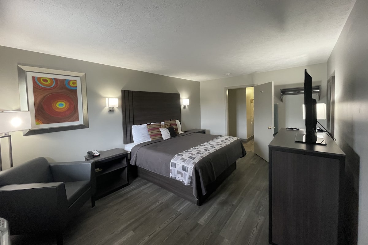 A hotel room with a bed, bedside tables, a desk, a chair, a TV, and a painting on the wall.