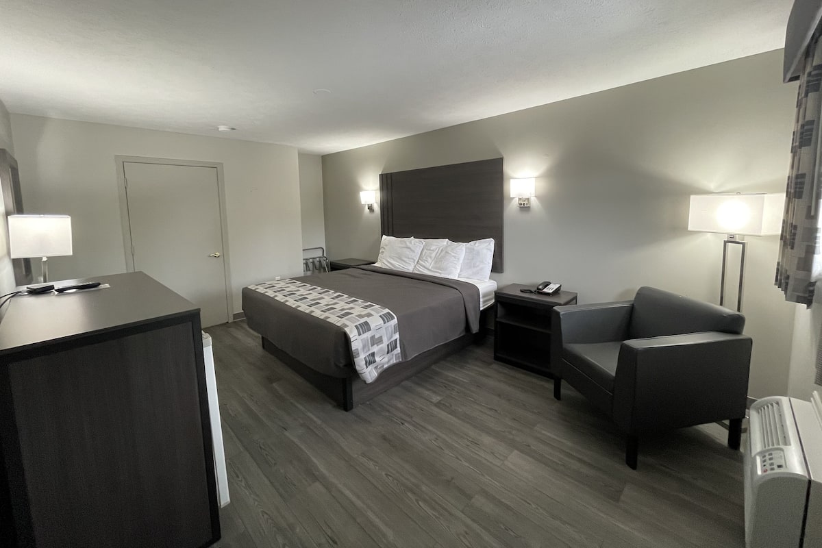 A hotel room with a bed, bedside table, chair, desk, and a heater.
